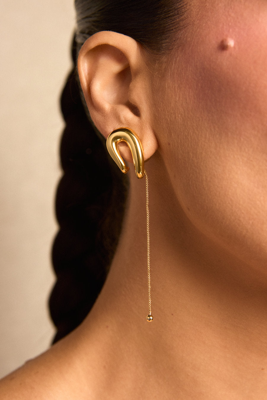 A close-up of a model's ear wearing a gold crescent-shaped dangle earring with a long chain and small bead at the end, against a warm beige background.