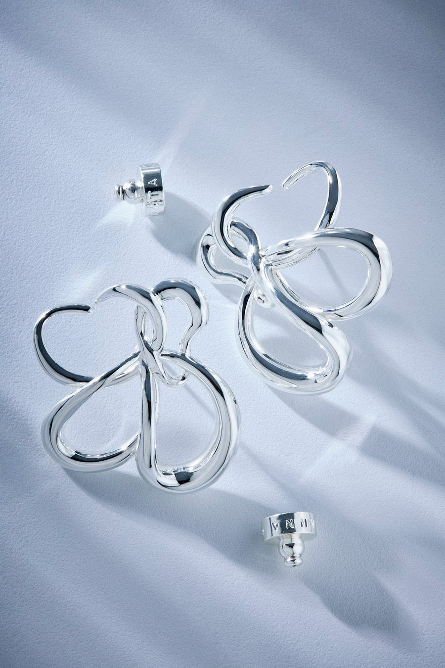 A pair of polished silver openwork earrings with a sculptural, flowing loop design, displayed on a light blue-gray textured fabric background with three visible post-backs.