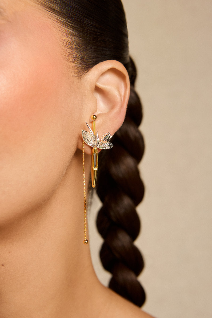 A close-up of a model's ear wearing a gold hoop earring with a dangling chain and a silver-toned dragonfly charm embellished with clear crystals, against a neutral beige background.