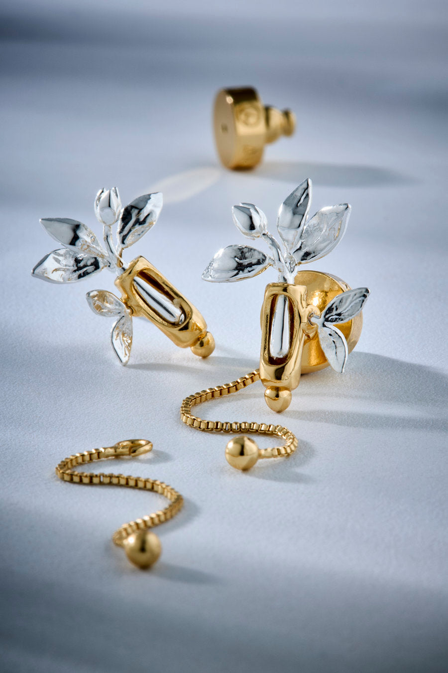 A pair of gold and silver leaf-shaped stud earrings with a matching gold beaded chain and ball, displayed on a light gray surface with a soft shadow.