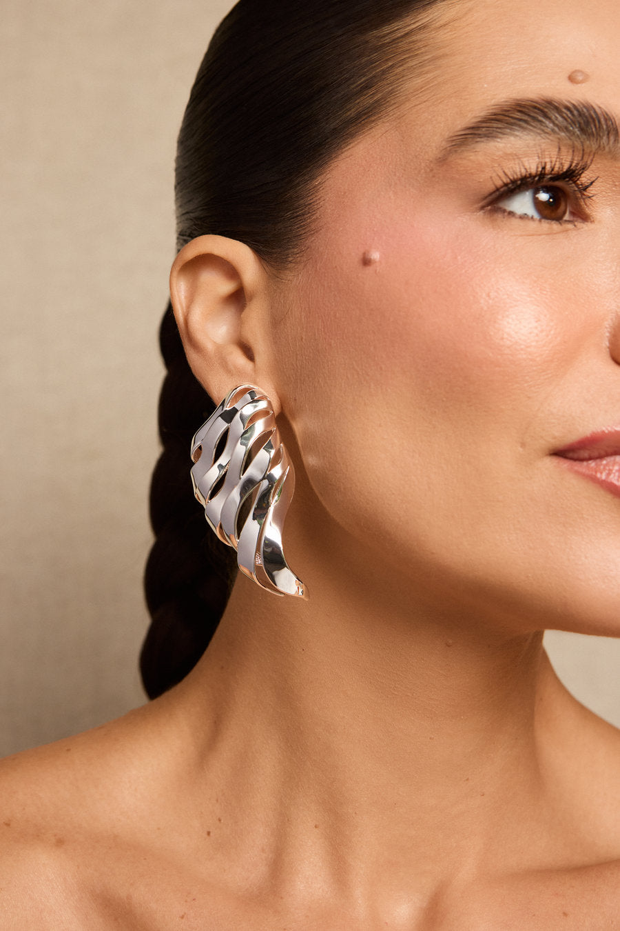 A close-up of a woman wearing a large, sculptural, polished silver earring with a wavy, layered design.
