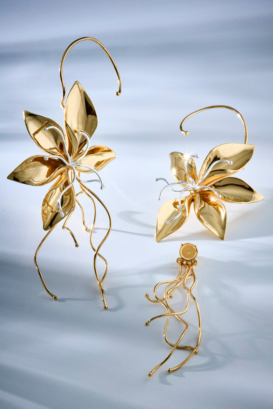 Three gold-toned floral hoop earrings displayed on a soft light blue fabric background. The set includes two larger flower-shaped hoops with long dangling wire elements and one smaller matching hoop.