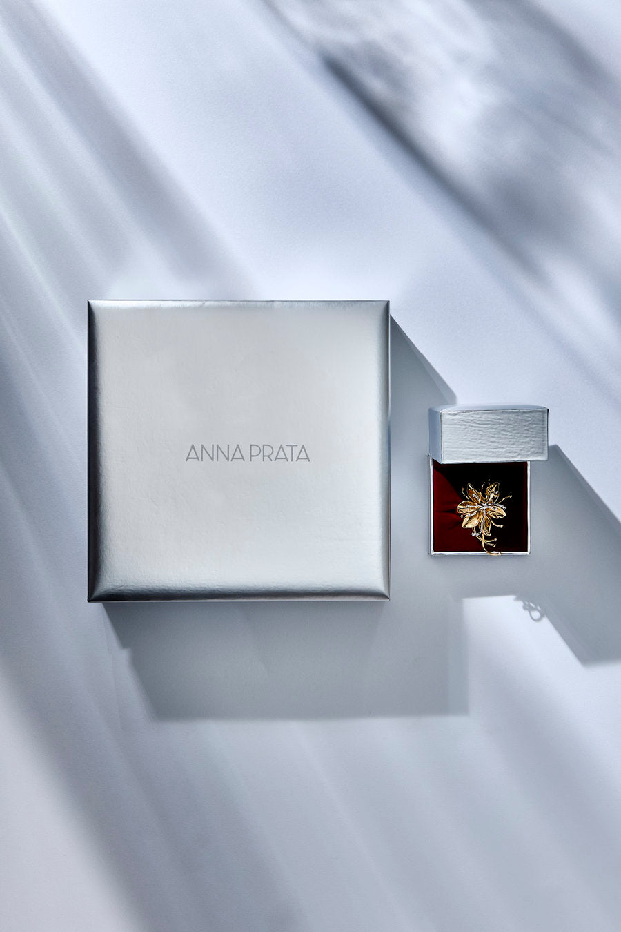 Anna Prata jewelry packaging and product shown on a draped light gray fabric background. A large square silver box with the brand name 'ANNA PRATA' is beside an open silver jewelry box containing a gold-toned brooch with a red insert.