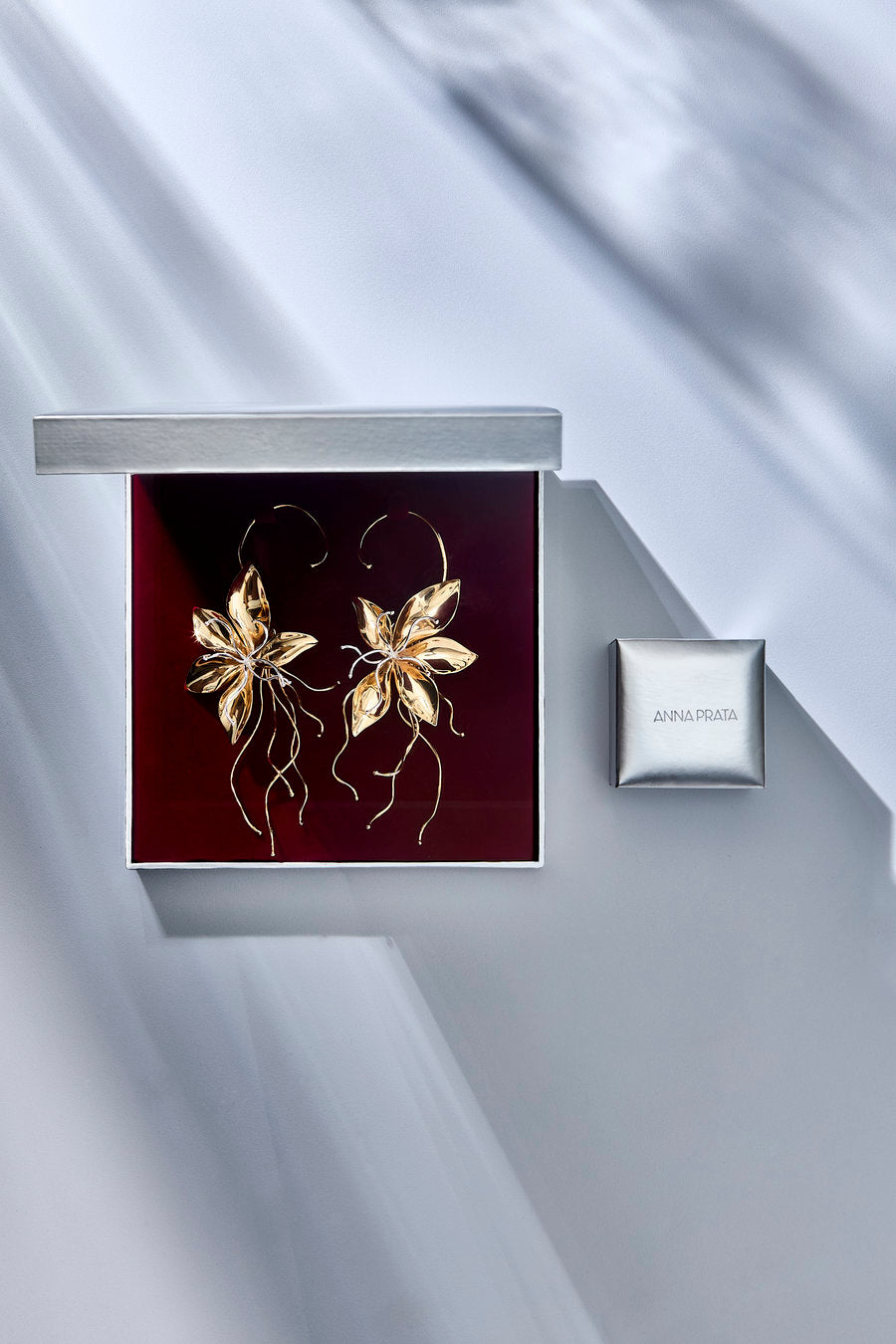 A pair of gold-toned floral hoop earrings displayed inside an open square box with a deep burgundy insert, shown beside a small branded silver box on a light gray draped surface.