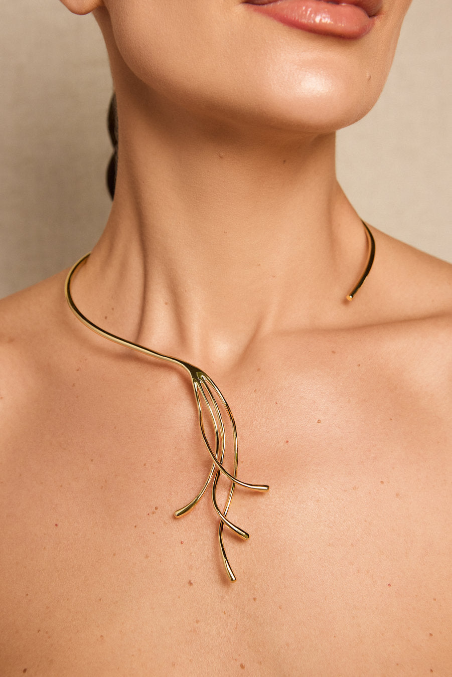 A close-up of a model's neck and upper chest wearing a delicate gold necklace with a branching, organic wire design.