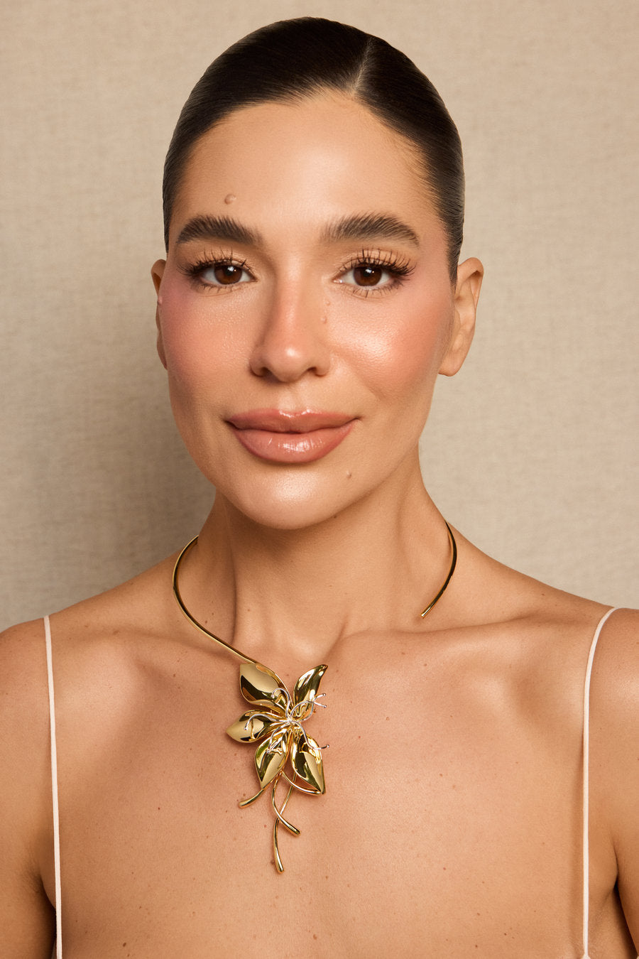A close-up portrait of a woman wearing a gold floral pendant necklace against a neutral beige background.