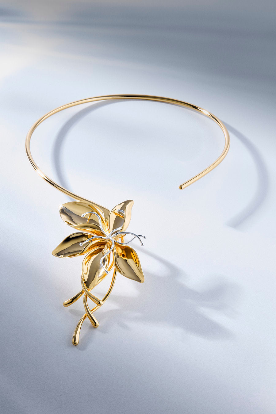 A polished gold-tone metal headband featuring a sculptural, leaf-like flower design on the front, displayed on a clean, light gray gradient background.