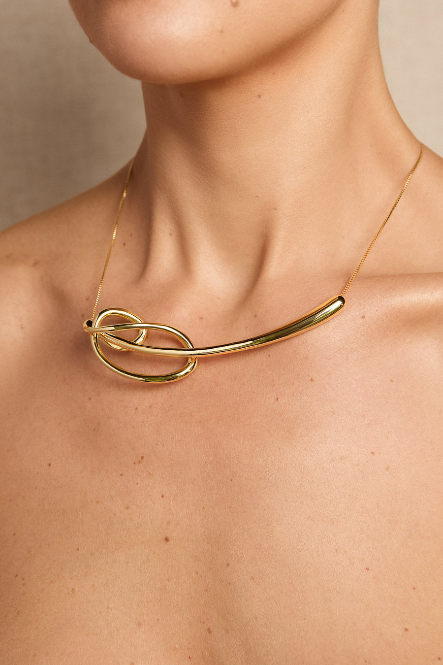 A close-up of a model's neck and upper chest wearing a polished gold necklace with a curved bar and interlocking loop design.