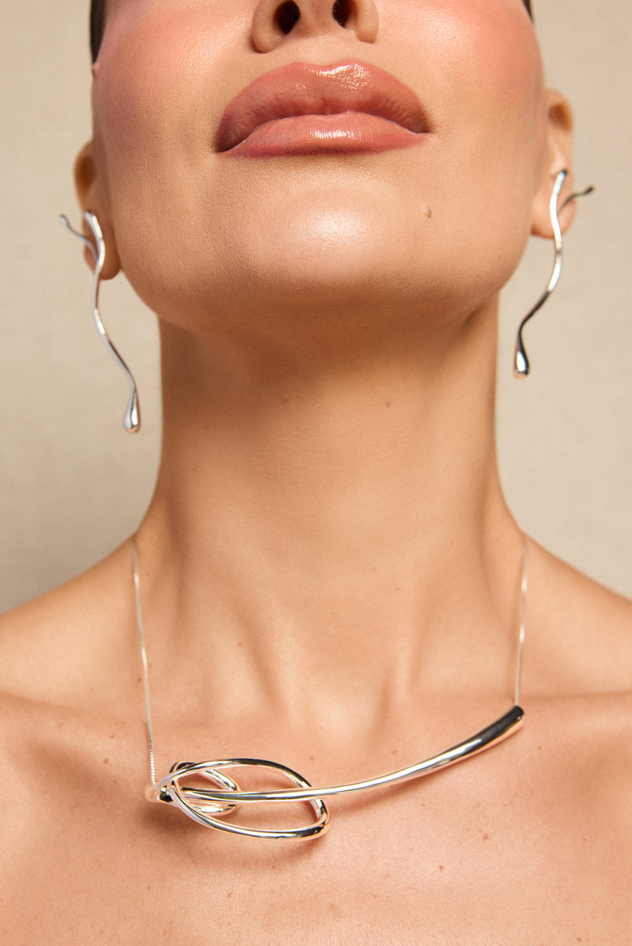A close-up of a model wearing a polished silver necklace with a curved bar and interlocking loop design, along with matching wavy hoop earrings, against a neutral beige background.