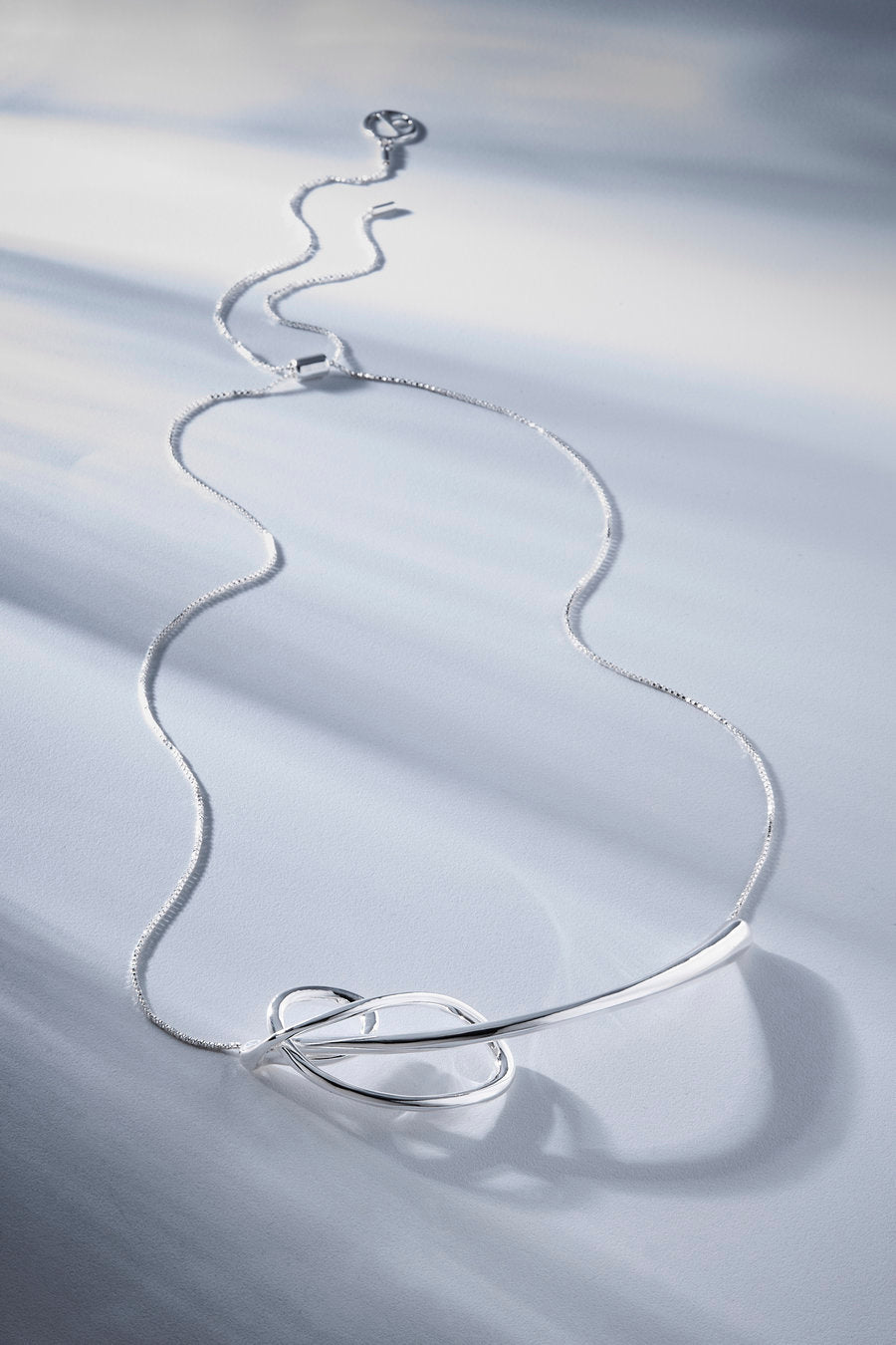 A silver snake chain necklace with a polished barbell pendant, arranged in a loose loop on a white textured fabric background with soft shadows.